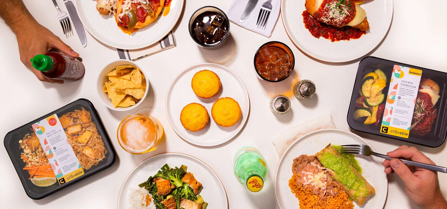Overhead tablescape of Craveble ready-to-eat trays, including pad thai with tofu and chicken parmesan, among plated dishes, drinks and cornbread, with hands reaching in.
