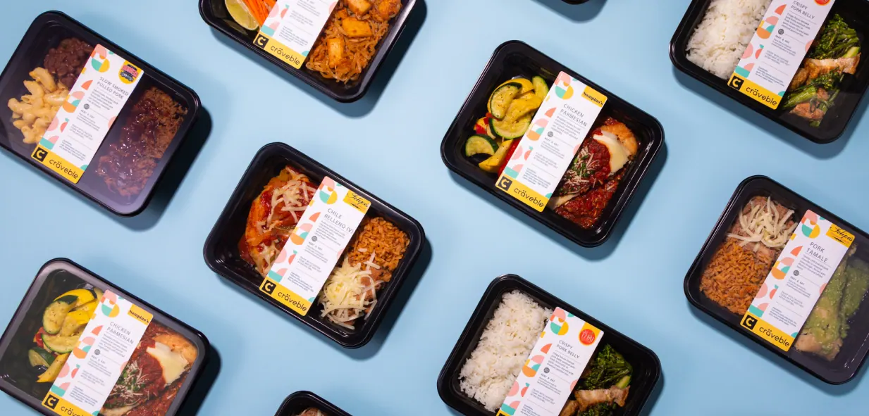Overhead flat-lay of sealed Craveble meal trays, including chicken parmesan, chile relleno and slow-smoked pulled pork, set diagonally on powder blue.