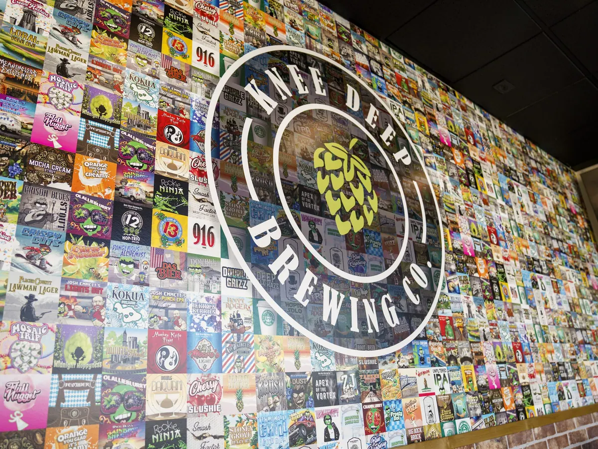 Knee Deep Brewing's Folsom taphouse wall, tiled floor to ceiling with colorful beer labels under a large circular Knee Deep Brewing Co. logo.