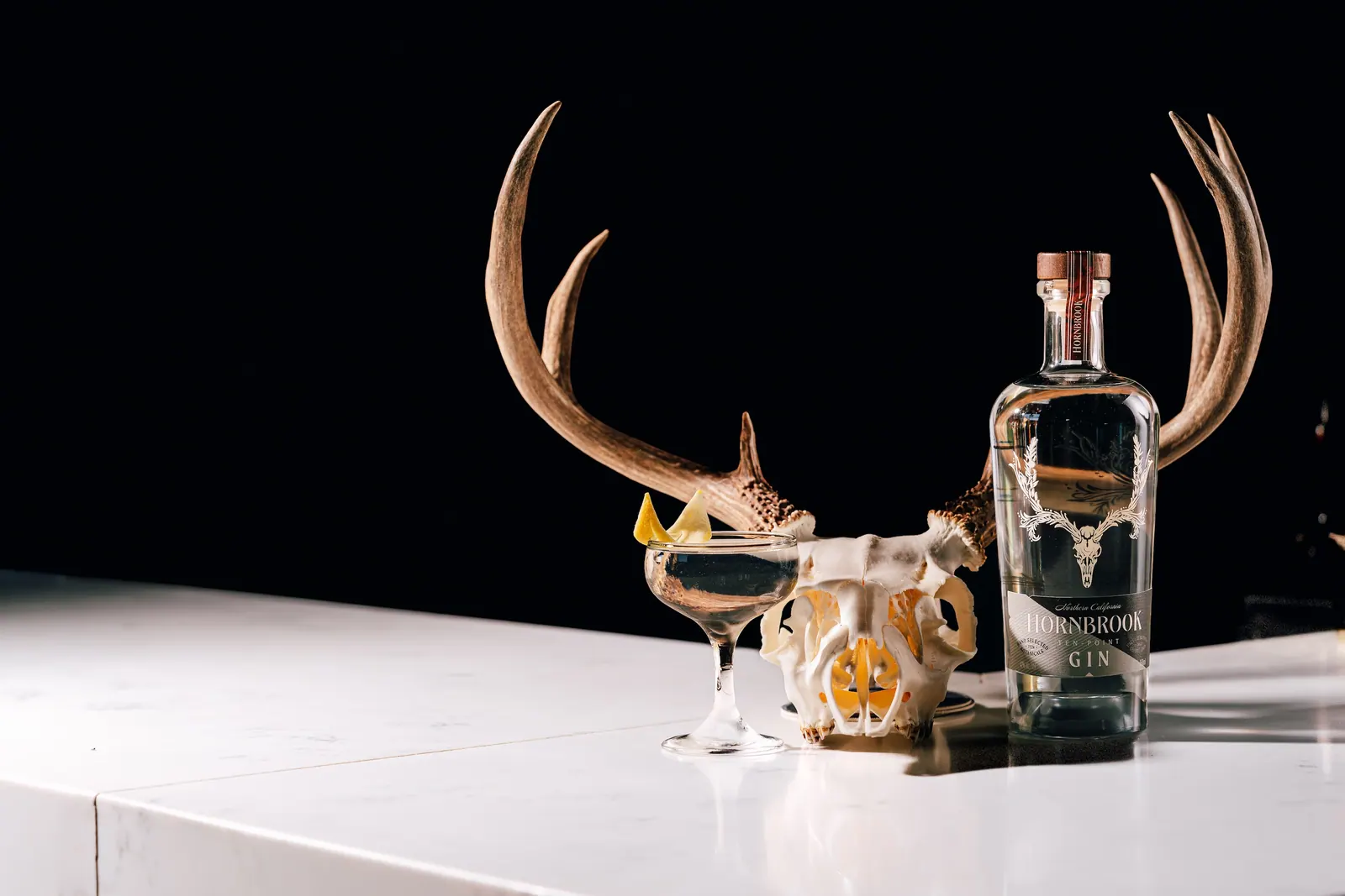 A bottle of Hornbrook Ten-Point Gin beside a coupe cocktail and a stag skull with antlers, on white marble against a black backdrop.